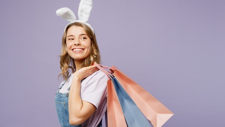 Lächelnde Person mit weißen Plüschohren steht mit einigen Präsenttaschen und Jeans-Latzhose vor einem lila Hintergrund