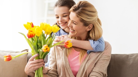 Eine Person sitzt auf einem Sofa und hält in der rechten Hand einen Blumenstrauß aus Tulpen. Die Person wird von hinten von einem Kind umarmt.