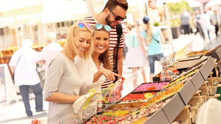 Drei Personen mit Sonnenbrillen stehen an einem Stand. Auf dem Stand sind in mehreren Behältern verschiedene Süßigkeiten. Eine der Personen deutet auf die Süßigkeiten. Im Hintergrund sind weitere Stände und Personen