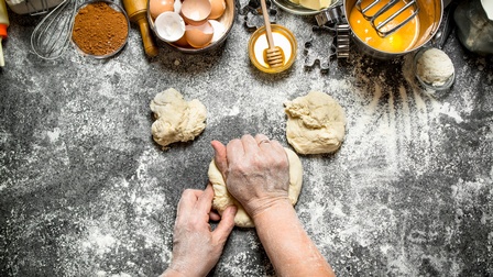 Topshot einer Person die einen Teig auf einer bemehlten Oberfläche knetet. Daneben liegen zwei weitere, kleine Teigstücke. Neben den Teiglingen sind Eierschalen, Honig, Butter, Keksausstecher, ein Schneebesen, Mehl sowie weitere Utensilien.
