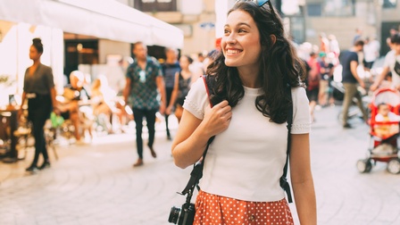 Eine Person mit hellem Oberteil, Rock, Sonnenbrille in den Haaren und Fotokamera um die rechte Schulter geht durch eine Fußgängerzone. Im Hintergrund sind weitere Personen sowie Außenbereiche von Lokalen