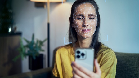 Person sitzt auf einer Couch in einem Wohnraum und hält ein Smartphone vor das Gesicht, auf der Gesichtsfläche sowie rund um das Gesicht zeigt sich eine Vermessung als Visualisierung der Gesichtserkennung