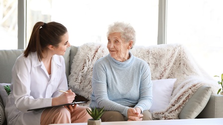 Person in weißer Arbeitskleidung sitzt bei einer betagten Person auf einer Couch und hält ein Klemmbrett sowie einen Stift in der Hand, im Hintergrund zeigt sich ein Wohnraum