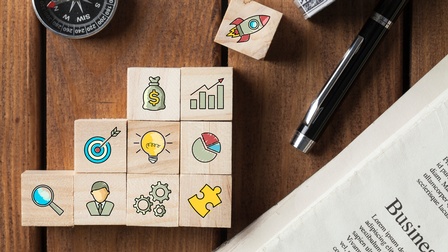 Auf einem braunen Holz-Untergrund liegt eine Zeitung, daneben ein Kugelschreiber und in der linken Bildhälfte ein Kompass. Mittig sind Holzwürfel zu Treppen gebaut. Darauf sind Symbole wie eine Rakete, Geldbeutel, Diagramme, eine Glühbirne, eine Lupe