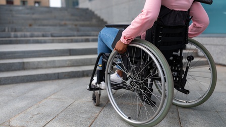 Person im Rollstuhl bewegt Räder mit Händen, davor ist eine Steintreppe ohne barrierefreiem Aufgang