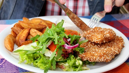 Nahaufnahme eines Schnitzels mit Sesam, Kartoffelspalten, Salt mit Gurken und Tomate sowie zweier Saucen auf einem Teller. Eine Person schneidet das Schnitzel<br />
