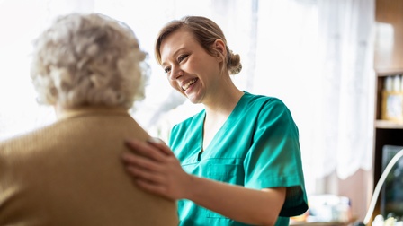 Lächelnde Person in grünem Arbeitshemd blickt auf ältere Person in Rückenansicht und berührt deren Schulter