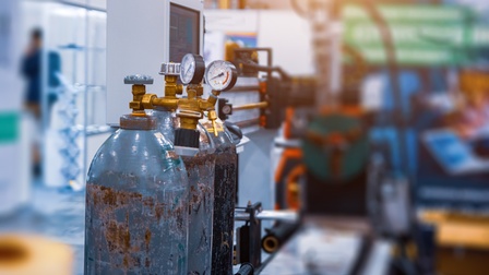 Im Fokus schwarze und blaue Gasflaschen mit Nutzungsspuren und Anlagenmonitor, im Hintergrund verschwommen Werkhalle und Person