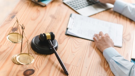 Detailansicht eines Schreibtischs mit Waage und Richterhammer, im Hintergrund verschwommen Hände einer Person, die auf Clipboard und Laptop liegen