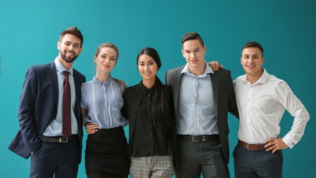 Porträt von fünf Personen in Bürokleidung, die nebeneinander vor einer türkisen Wand stehen. Die Personen haben die Arme umeinander gelegt und lächeln