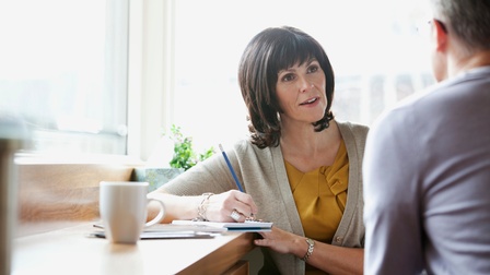Zwei Personen sitzen sich an einem Tresen gegenüber. Eine Person sitzt mit dem Rücken zur Kamera. Die andere Person notiert etwas auf einem Notizblock. Auf dem Tresen steht eine Tasse
