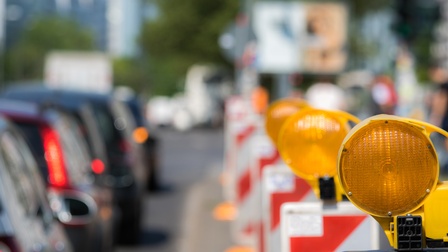 In der linken Bildhälfte sind mehrere hintereinander stehende Autos. In der rechten Bildhälfte sind viele hintereinander stehende rot-weiße Absperrungen. Auf jeder Absperrung ist oben ein oranges Licht montiert.
