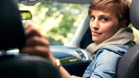 Person mit kurzen braunen Haaren, Schal und Jeansjacke sitzt auf der Beifahrerseite eines Autos und blickt auf die Rückbank zurück in die Kamera