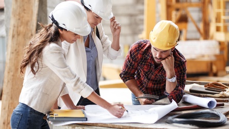 Drei Personen stehen um einen Bauplan. Alle drei tragen einen Helm. Die Person ganz links deutet mit einem Stift darauf