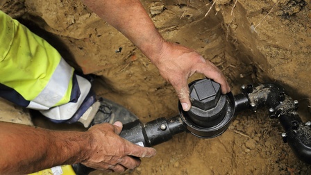 In einem Loch in der Erde verläuft ein schwarzes Rohr parallel zum Boden. Daneben steht eine Person. Die Person hat ihre beiden Hände auf dem Rohr