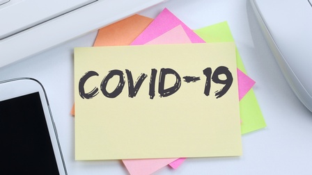 Verschiedenfarbige Post-its aufeinander gestapelt, am obersten der Schriftzug Covid-19, daneben im Ausschnitt Smartphone, Computermaus und Tastatur 