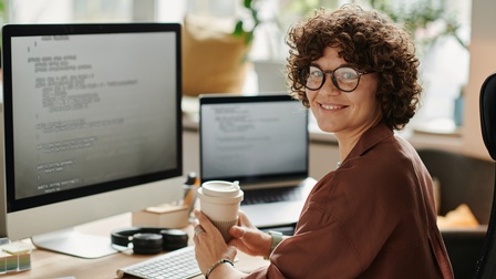 Eine zur Kamera gewandte Person mit Brille und kurzen, lockigen Haaren sitzt an einem Schreibtisch. Vor ihr steht ein Bildschirm und eine Tastatur. Rechts daneben steht ein aufgeklappter Laptop. In den Händen hält sie einen Papierbecher