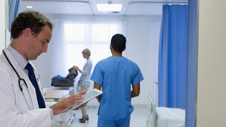 Seitliche Aufnahme einer Person in einem weißen Kittel, mit Stethoskop und mit Tablet in der Hand, auf das sie blickt. Im Hintergrund geht eine Person in blauer Kleidung sowie packt eine andere Person einen Koffer aus