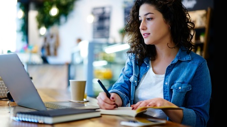 Person blickt auf aufgeklapptes Notebooks und nimmt mit Stift Notizen in Buch vor