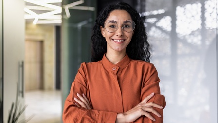Person mit dunklem lockigen Haar und Brille sowie oranger Bluse und verschränkten Armen blickt freudig in die Kamera, im Hintergrund zeigt sich ein moderner Raum