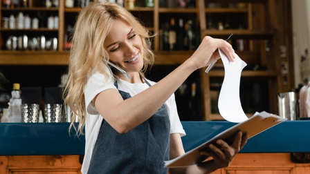 Person mit langen blonden Haaren und Schürze telefoniert mit einem Smartphone in der Schulter eingeklemmt und steht vor einer Schank und blickt auf Dokumente auf einem Klemmbrett
