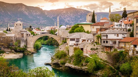 Über einen Fluss führt eine hohe Brücke aus Stein. Links und rechts davon sind leicht abfallend mehrere verschiedene Gebäude. Manche sind aus Stein. Im Hintergrund ist eine Bergkette