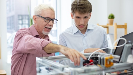 Person mit kurzen weißen Haaren, Bart, schwarzer Brille und rosa Hemd steht gemeinsam mit einer jugendlichen Person mit hellblauem Hemd und Laptop vor einem 3D-Drucker und begutachtet ihn