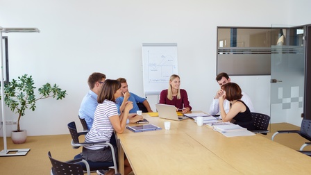 Personen sitzen rund um einen Besprechungstisch in einem Raum, vorne steht ein Flipchart mit einer aufgezeichneten Grafik