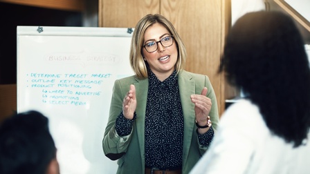 Eine Person in Bluse, Blazer und Brille steht vor einer weißen, beschrifteten Tafel. Ihr gegenüber sind zwei weitere Personen