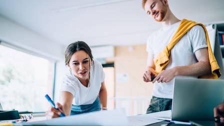 Person streckt sich über Tisch und markiert etwas mit Stift auf großem Papierbogen, andere Person sieht ihr mit geneigtem Kopf zu