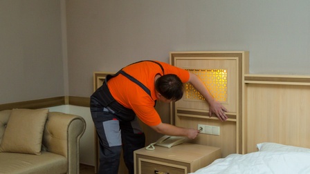 Eine Person in Latzhose steht neben einer Wand, beugt sich nach vorne und hält die rechte Hand vor eine weiße Steckdose, die über einem Kästchen ist. Das Kästchen steht neben einem Bett.