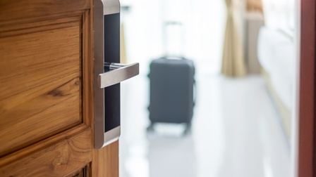 Tür eines Hotelzimmers ist geöffnet und im Zimmer steht in der Unschärfe ein Rollkoffer