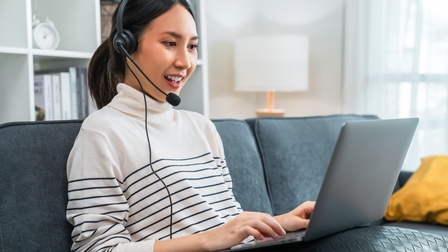 Sprechende Person mit Headset in gestreiftem Pullover sitzt auf petrolfarbener Couch und blickt auf vor ihr am Schoß aufgeklappten Laptop, im Hintergrund weißes Bücherregal mit Büchern und Wecker, nebenstehend weiße Tischlampe