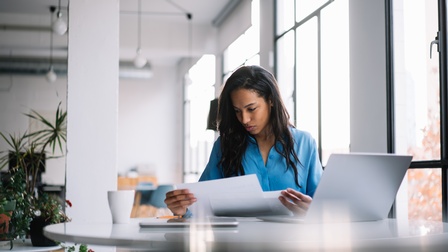 Person sitzt in hellem Büroraum vor Laptop und sichtet Dokumente