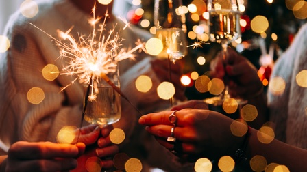 Nahaufnahme der Hände von drei Personen, die in einem Kreis stehen. Jede Person hält in einer Hand ein Sektglas mit Sekt und in der anderen Hand eine Wunderkerze