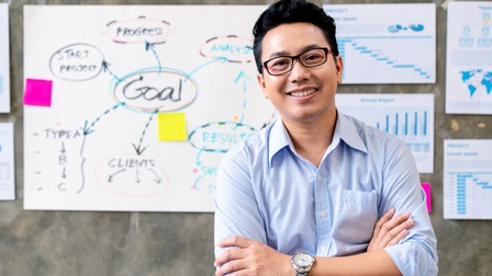 Halbporträt einer Person mit kurzen, dunklen Haaren und verschränkten Armen. Hinter der Person hängt an einer Wand ein Plakat, auf dem handschriftlich Begriffe zum Thema Strategie steht. Rechts daneben sind bedruckte Zettel mit Grafiken an der Wand