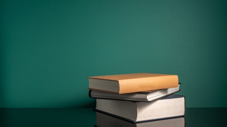 Stapel aus drei Hardcoverbüchern, der sich auf darunterliegendem Untergrund spiegelt vor grüner Wand