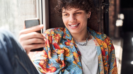 Eine jugendliche Person mit kurzem, lockigem Haar, einer silbernen Kette und einem Nasenpiercing sitzt auf einem Fensterbrett und lehnt sich mit dem Rücken am Fensterrahmen an. In der rechten Hand hält sie ein Handy.