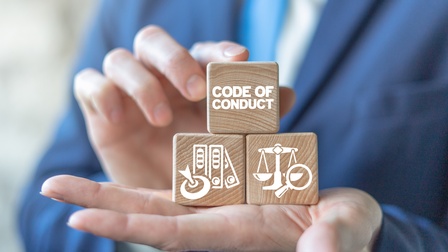 Die nach oben geöffnete Hand einer Person hält drei Holzquader. Auf dem obersten steht in weißer Schrift code of conduct. Auf dem links darunter sind Ordner und eine Zielscheibe. Auf dem daneben sind Symbole  einer Waage und eine Lupe