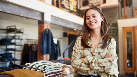 Halbporträt einer Person mit verschränkten Armen. Die Person steht in einem Raum. Hinter ihr führen Stiegen nach oben. Links neben ihr ist ein Tisch mit gestapelter, gefalteter Kleidung. Links an der Wand sind Kleiderständer mit Kleidungsstücken.