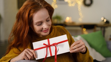 Auf dem Bild ist eine lächelnde Frau zu sehen, die einen Geschenkgutschein in der Hand hält. Im Hintergrund sieht man eine Couch und einen Schreibtisch. Darüber hängen Lichterketten.