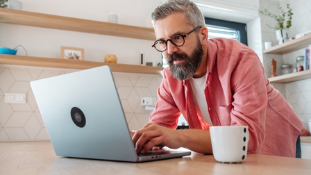 Eine Person mit grauem Bart und grauem Haar sowie mit Brille beugt sich über einen Tisch in einer Küche und tippt auf der Tastatur eines aufgeklappten Laptops. Daneben steht eine Tasse