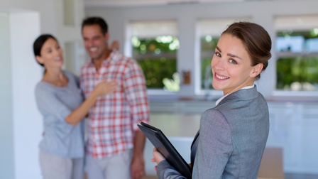 Eine Person in einem Sakko mit einer Unterlagenmappe dreht sich zur Kamera und lächelt. Ihr gegenüber stehen zwei Personen eng nebeneinander. Die Personen stehen in einem Wohnraum. Hinter ihnen stehen Kartons