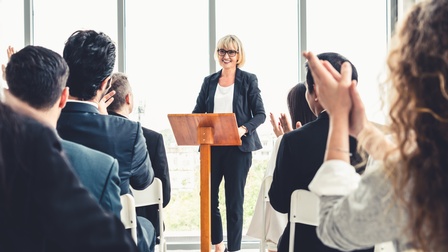Personen sitzen im Publikum mit dem Rücken zur Kamera, vorne steht eine vortragende Person mit dunklem Sakko, dunkler Hose, hellem Oberteil und Brille hinter einem Rednerpult