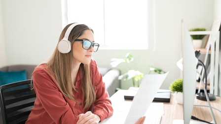 Seitliche Aufnahme einer Person mit langen, hellen, offenen Haaren, Brille sowie Kopfhörern, die an einem Schreibtisch sitzt und einen Zettel hält. Vor der Person steht ein Bildschirm
