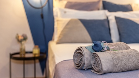 Fokus auf zusammengelegten Handtüchern auf frisch gemachtem Bett, neben dem Bett steht ein Beistelltisch mit frischen Blumen, eine Lampe erhellt den Raum