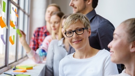Lächelnde Person mit Brillen im Fokus, umgeben von weiteren Personen die auf Fenster Post-its kleben und betrachten
