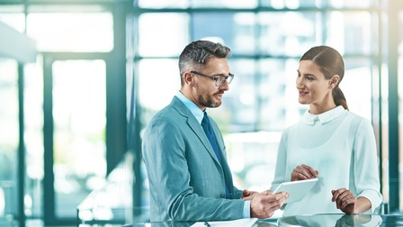 Zwei Personen stehen an Tischen in Unterhaltung, eine Person hält Tablet in Händen, am Tisch Dokument mit Stift liegend