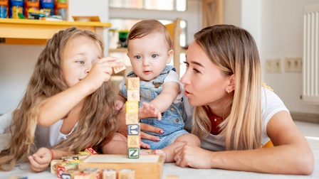 In der Mitte sitzt ein Kleinkind. Links daneben liegt ein Kind auf dem Bauch. Rechts neben dem Kleinkind ist eine erwachsene Person, die das Kleinkind mit dem rechten Arm umfasst. Vor ihnen steht ein Turm aus Holzwürfeln