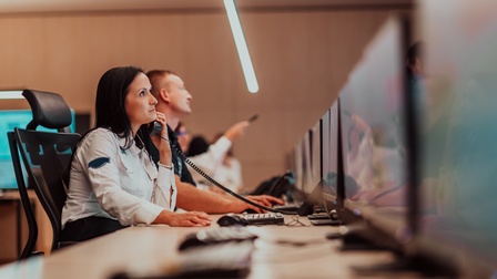 Person in Arbeitskleidung sitzt an einem Schreibtisch und telefoniert während daneben weitere Personen sitzen
